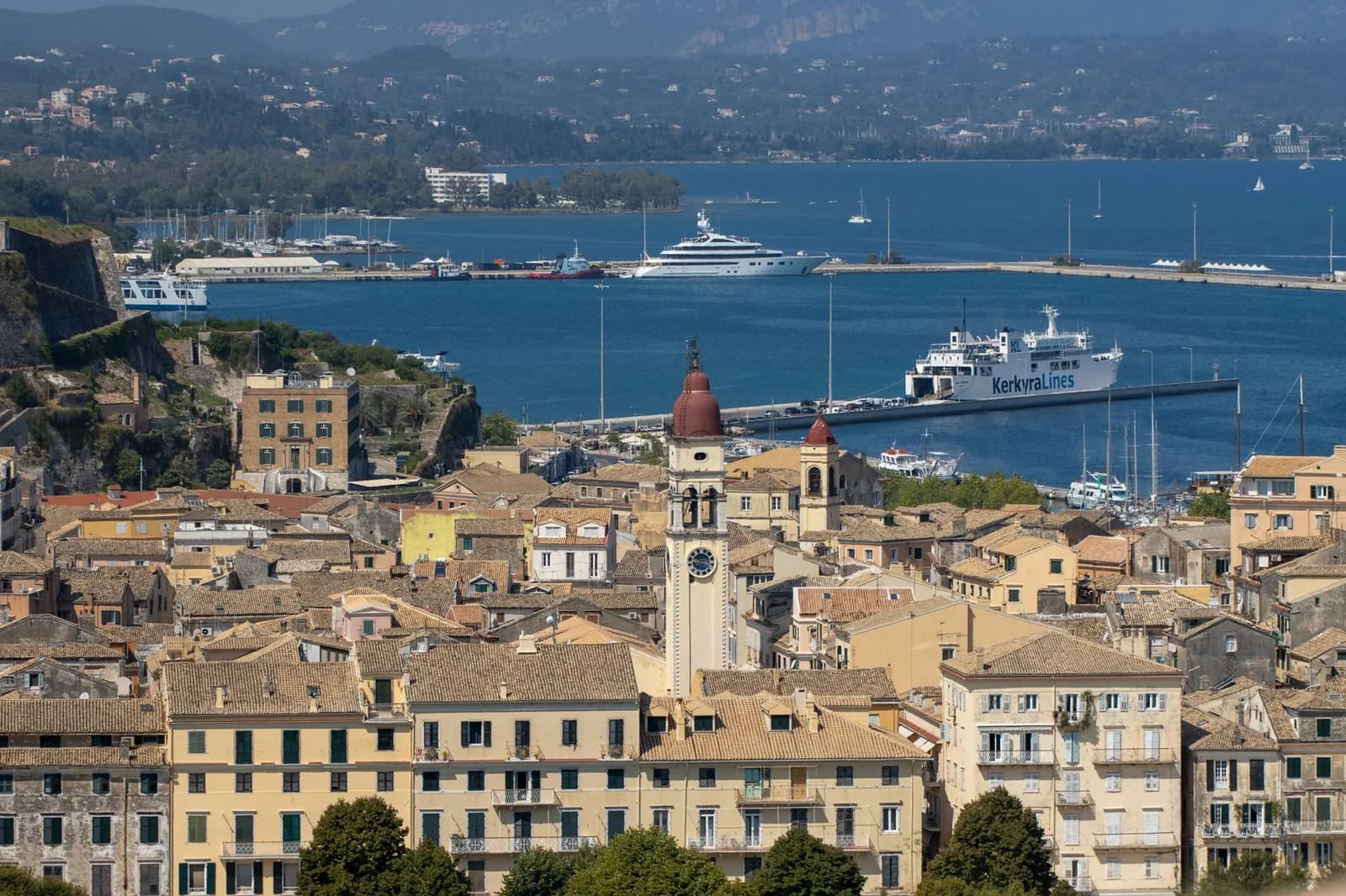 A KerkyraLines ferry berthed at the old port of Corfu town with cars boarding, the Venetian old town and the campanile of Saint Spyridon in front of it, and the mainland coast across the strait