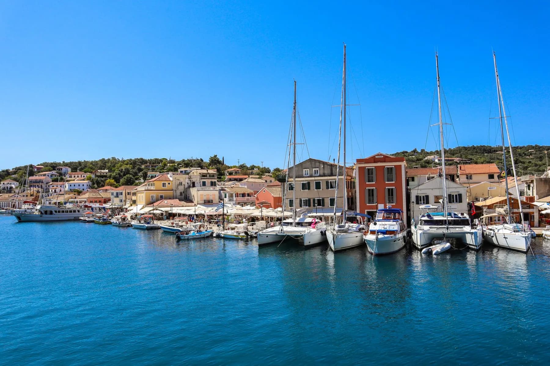 The waterfront at Gaios on Paxos, with pastel Venetian houses and café awnings along the quay, yachts and a small ferry moored in front and wooded green hills rising behind the village