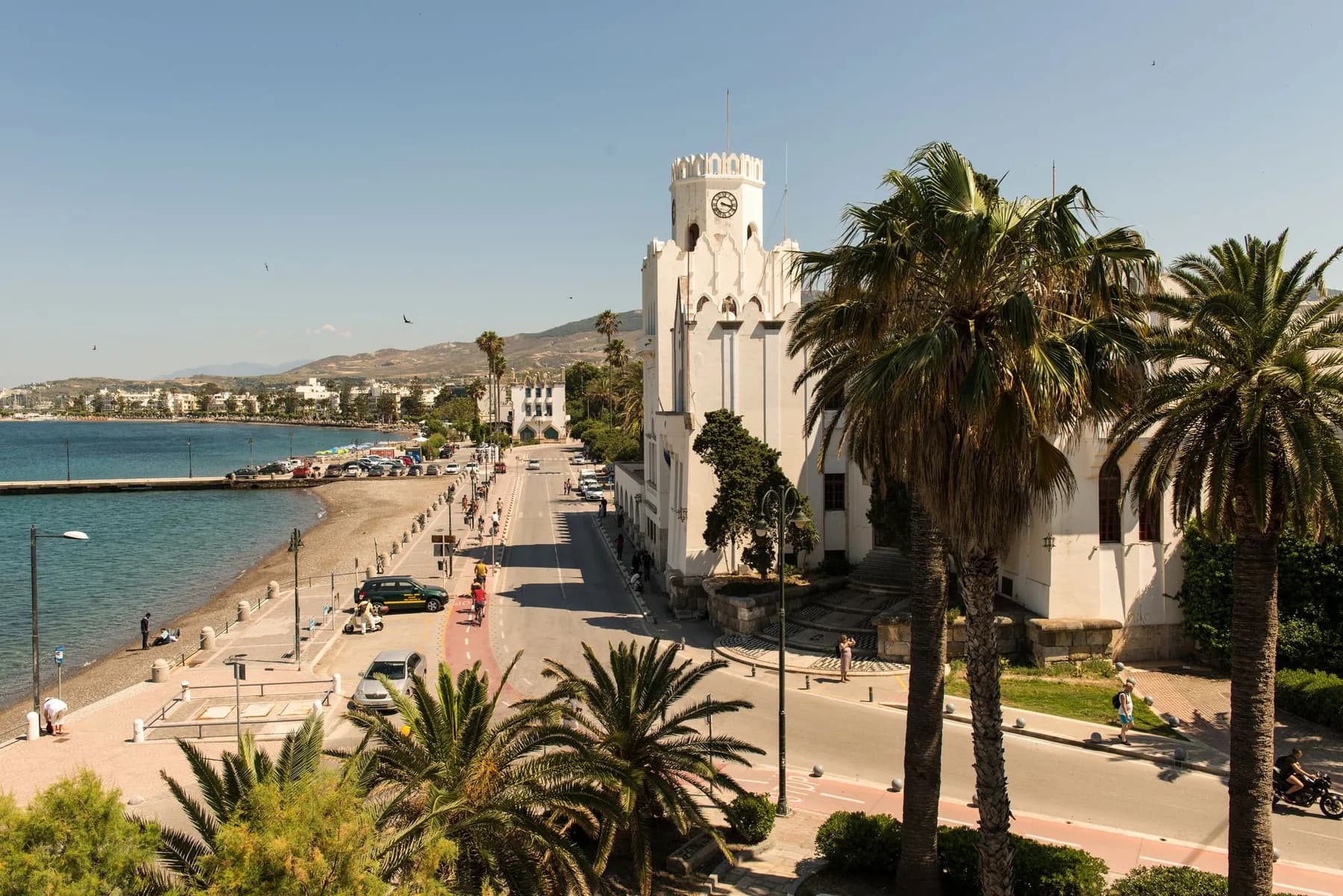 The seafront avenue in Kos Town with a marked red cycle lane, cyclists, palms and the Italian-built administrative building with its clock tower on the right