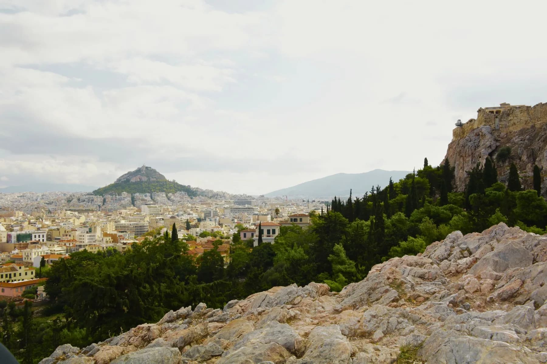 Lycabettus Hill rising as a wooded cone above the rooftops of central Athens, seen from the rock of the Acropolis with the fortification wall of the site on the right