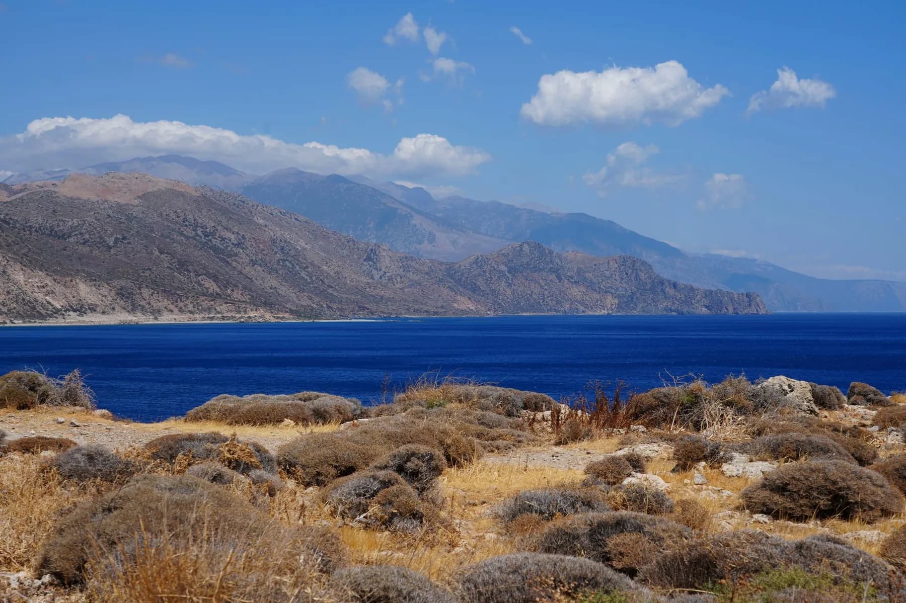 A range of bare mountains on Crete running along the coastline and dropping straight into deep blue sea, seen across scrub burnt brown by the summer