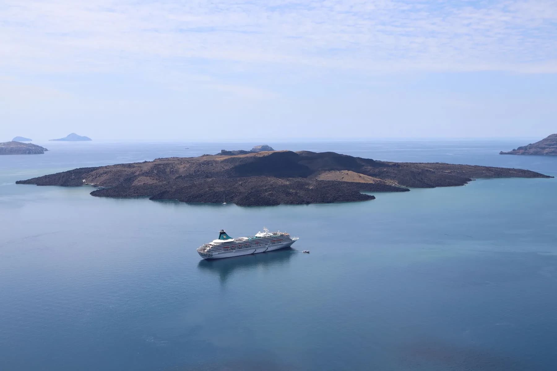The black lava island of Nea Kameni lying in the middle of the flooded caldera of Santorini, with a cruise ship anchored off it and the rim of the crater on both horizons