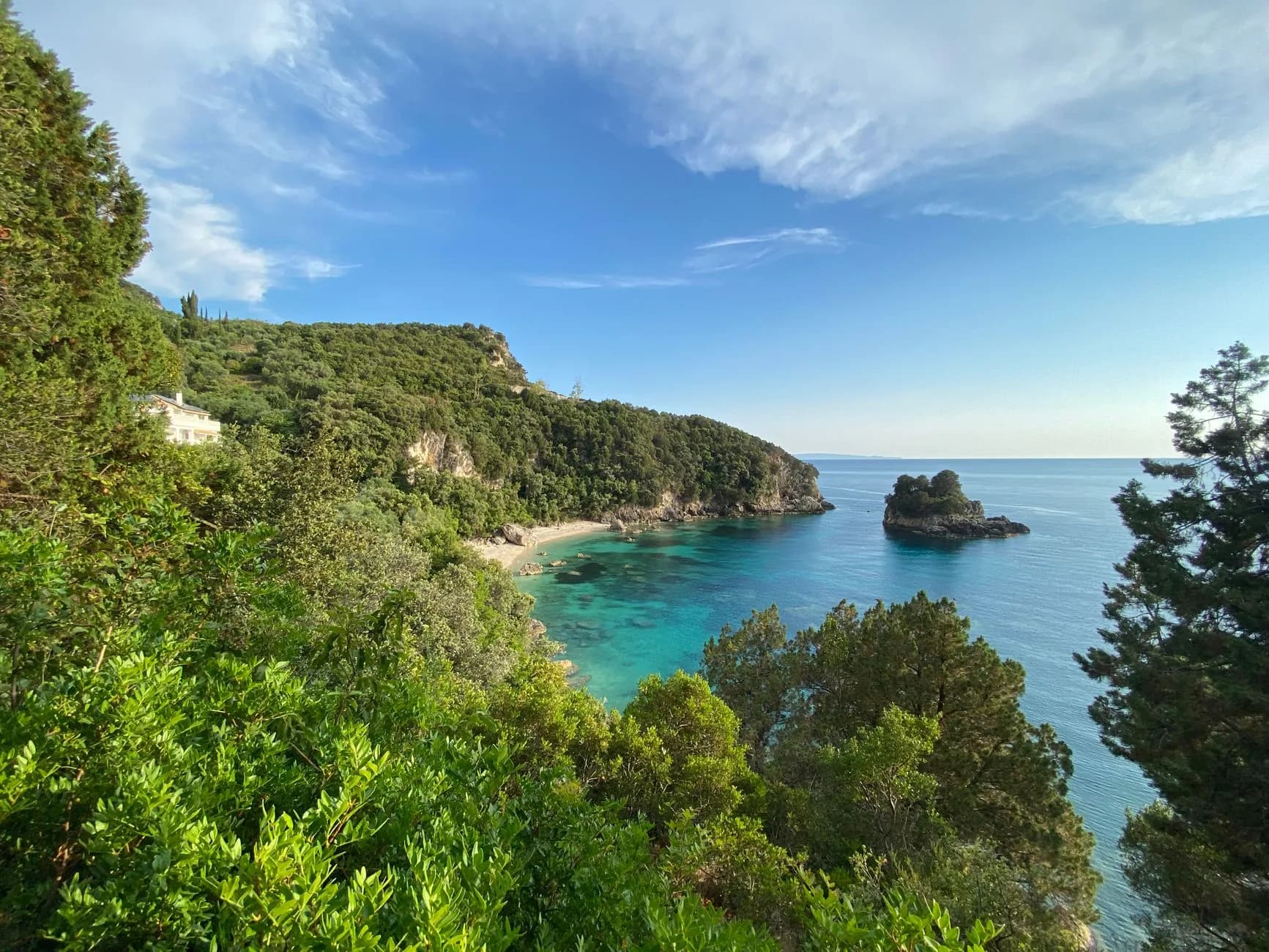 A densely wooded headland on the west coast of Corfu dropping into a turquoise cove, with a small rock islet offshore