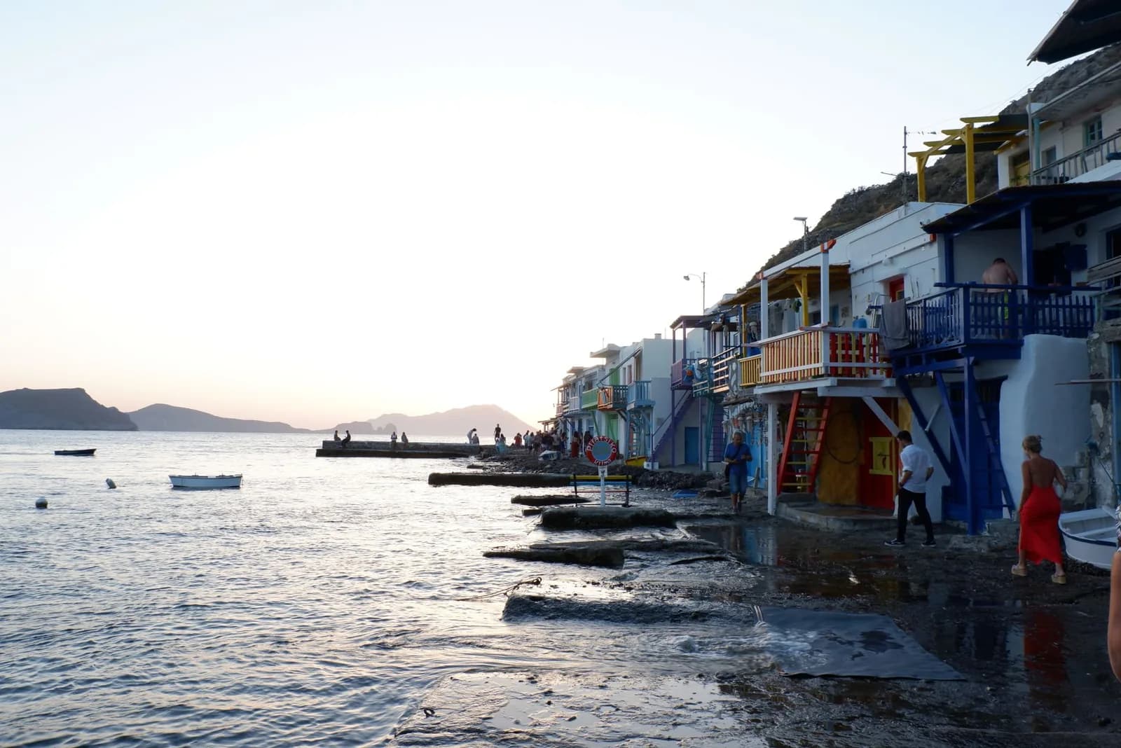 The waterfront row at Klima on Milos at sunset, two-storey fishermen's houses with balconies and doors painted red, yellow, blue and green standing directly on the shoreline, with headlands silhouetted across the water