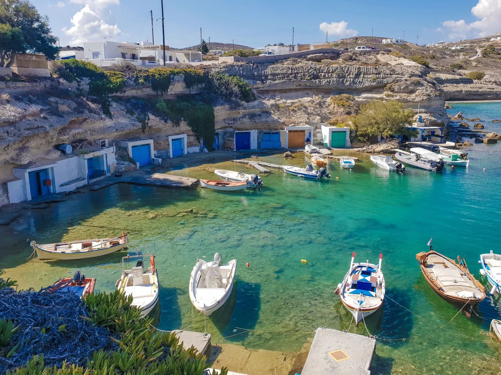 A sheltered fishing cove on Milos with a row of whitewashed boat garages built into the rock at the waterline, their doors painted blue, turquoise and orange, small fishing boats moored in clear green water and a low village on the ridge above