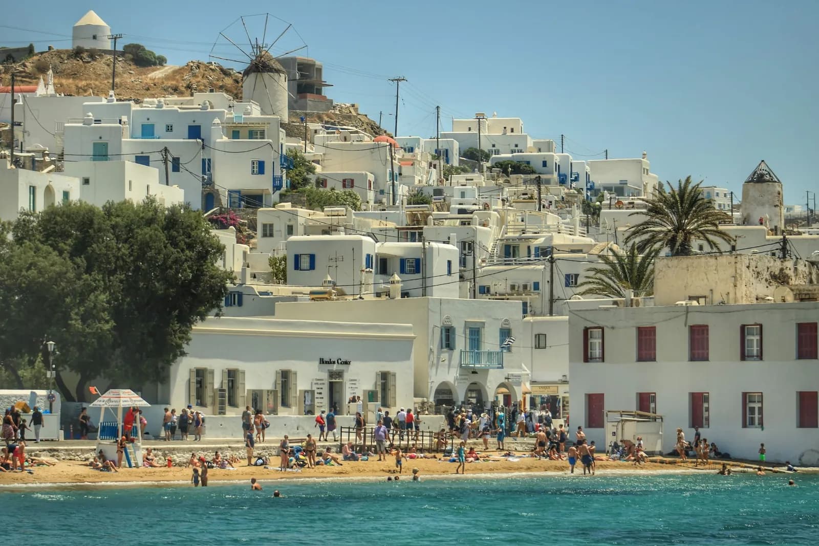 The whitewashed houses of Mykonos Chora stacked up the hillside above the waterfront, with a stone windmill on the ridge behind them and people on the small town beach in front