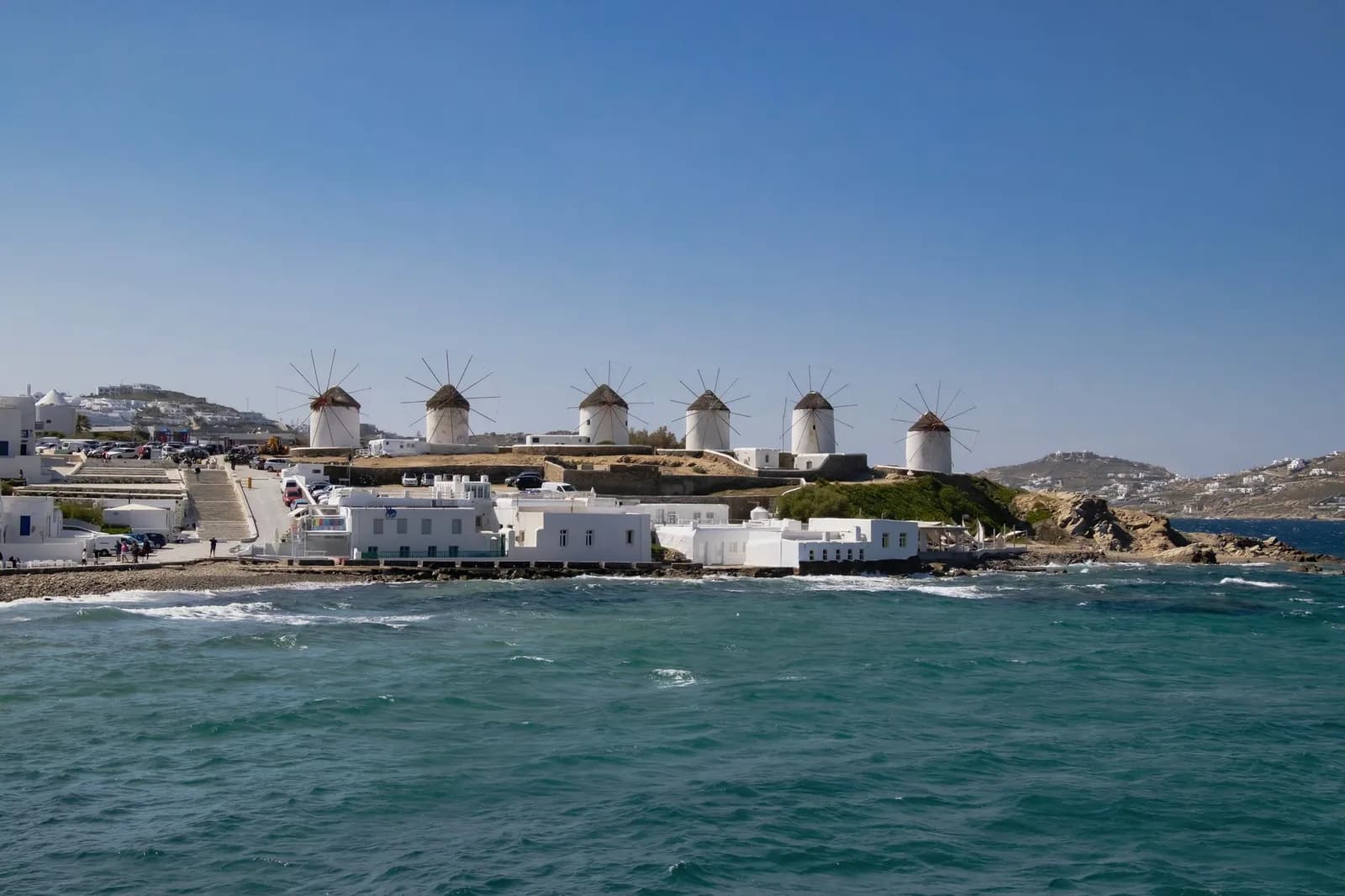 The row of Kato Mili windmills on the rise above Mykonos Chora, with the car park and coast road below them, seen from the water