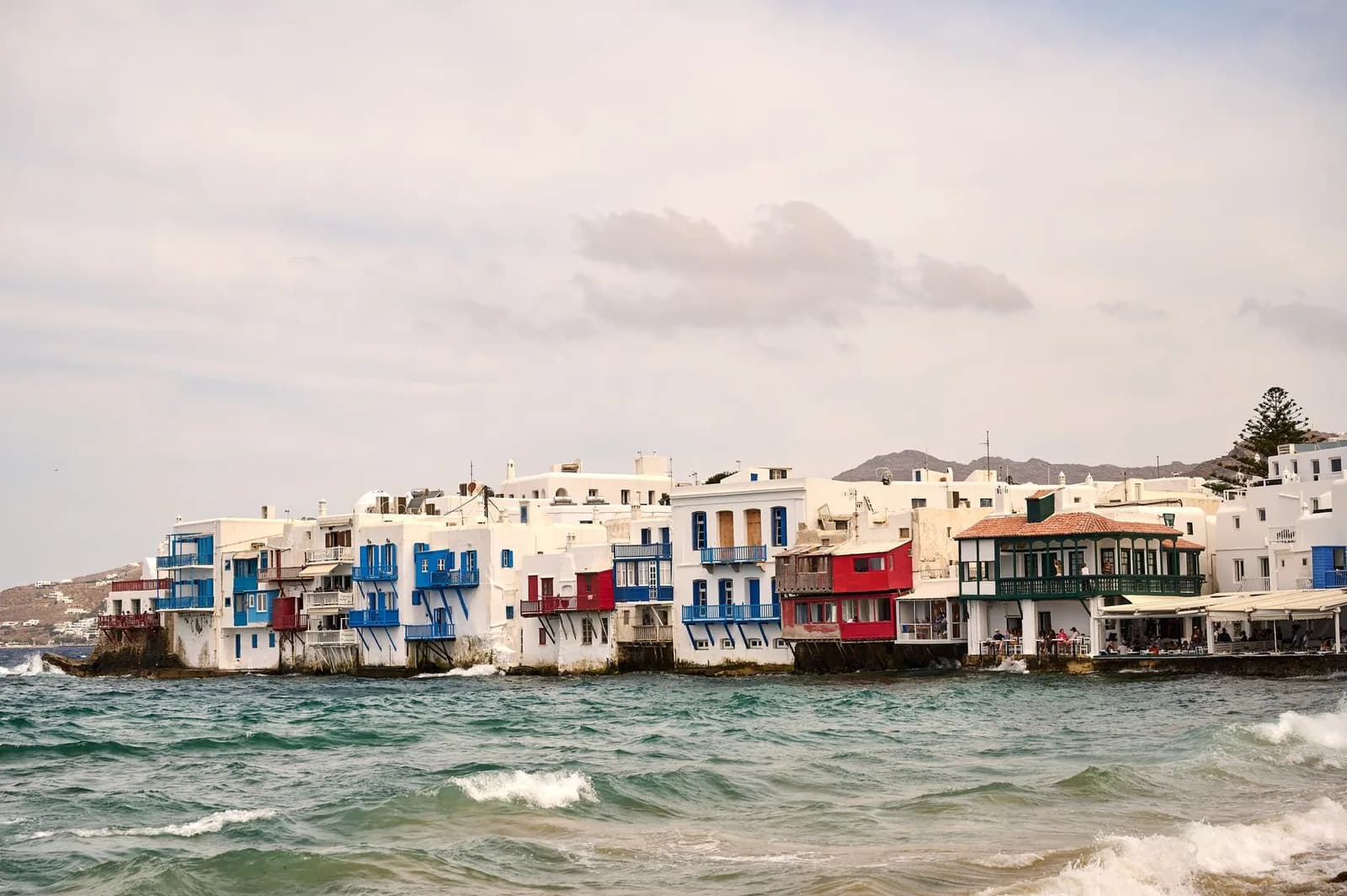 The row of houses at Little Venice on the western waterfront of Mykonos Chora, their blue and red wooden balconies built out over the sea with waves breaking against the foundations below them
