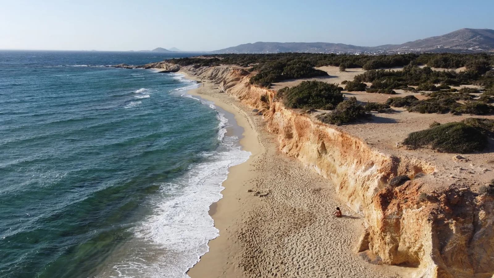 Aerial view of the southern west coast of Naxos, a long empty sand beach below ochre sandstone cliffs with cedar scrub and dunes behind it and the island's mountains in the distance