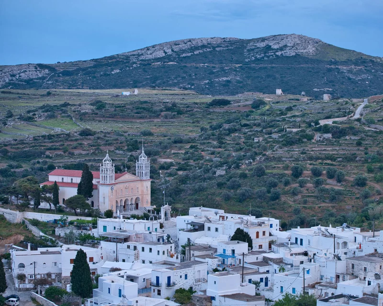 The village of Lefkes on Paros seen from above at dusk, with the twin marble bell towers of the church of Agia Triada above the whitewashed houses and the terraced flank of Profitis Ilias rising behind