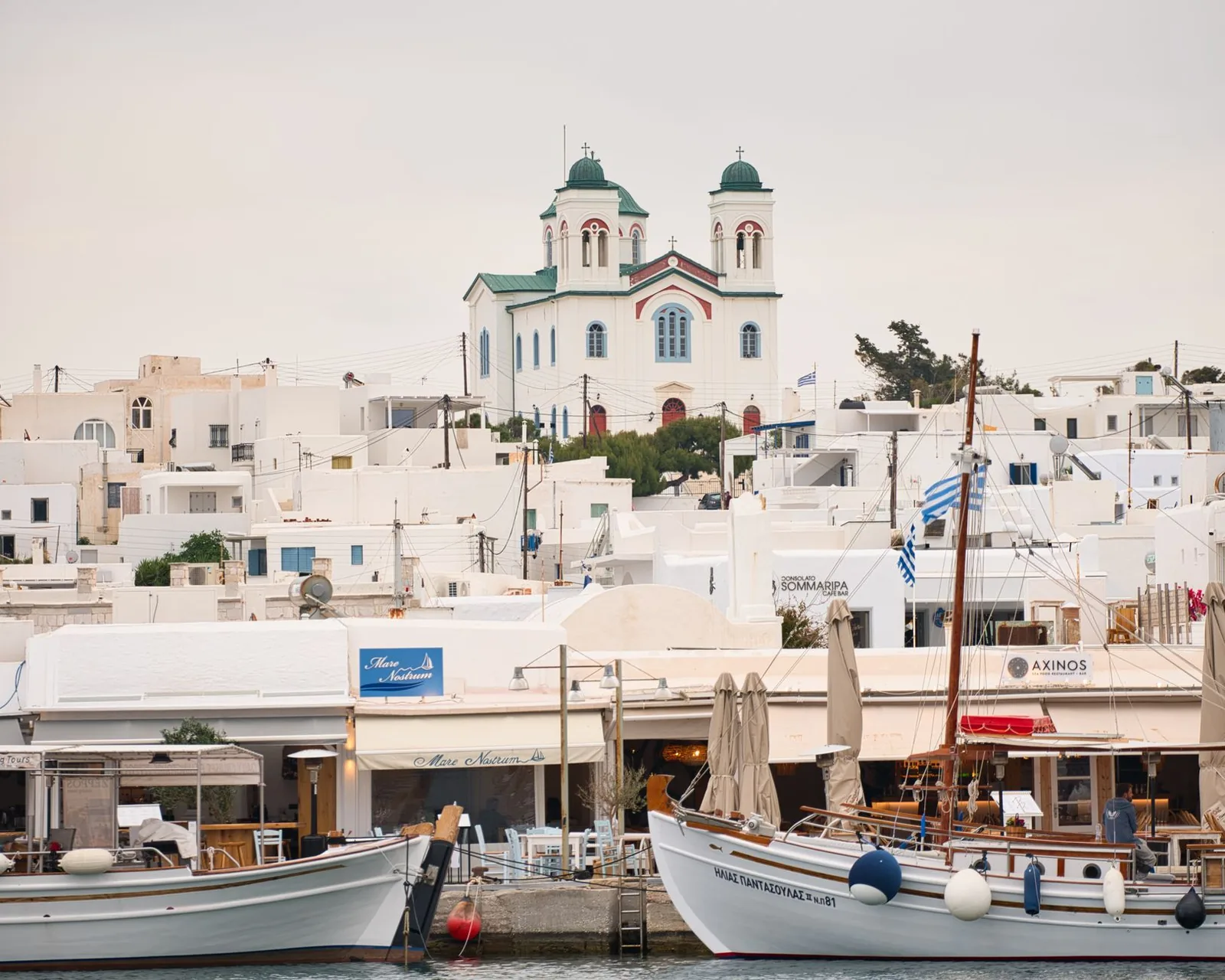 The fishing harbour of Naoussa on the north coast of Paros, with wooden caiques tied along the quay, tavernas on the waterfront and the twin bell towers of the church rising above the whitewashed town behind