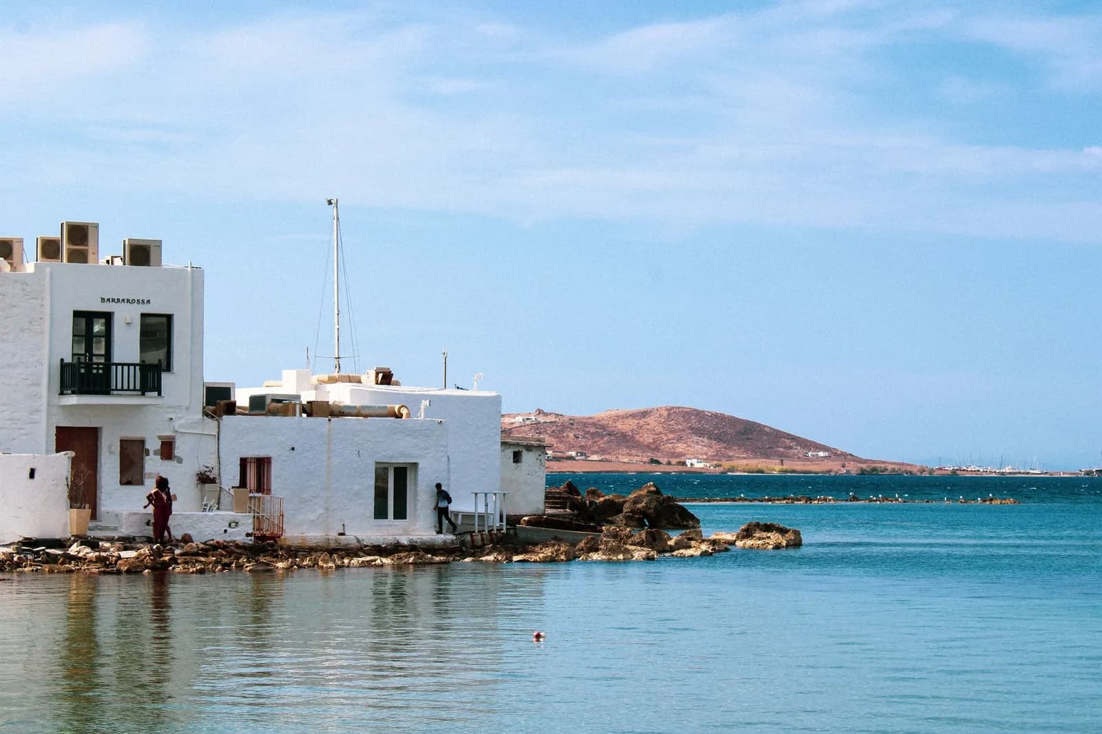 Whitewashed houses at the water's edge on the western side of Naoussa, Paros, with rocks in the shallows and the bare headland on the far side of the bay behind them