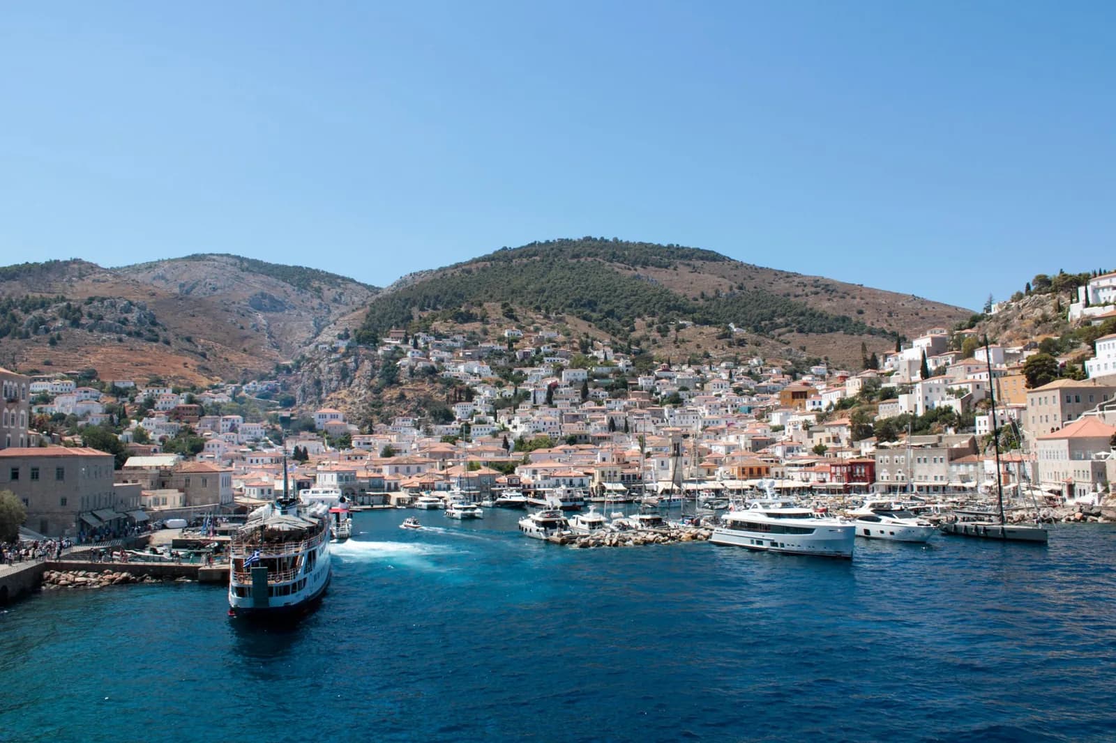 A passenger boat turning into Hydra harbour, with the stone houses of Hydra town climbing the hills behind